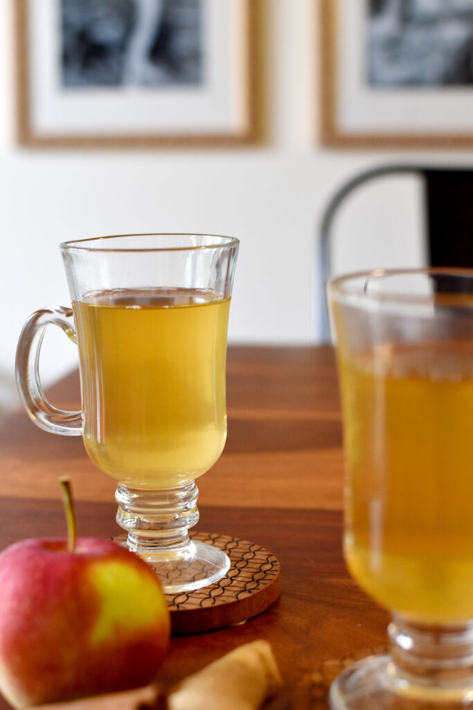 a lifestyle shot of two people enjoying cinnamon apple tea in cute glass irish coffee mugs with handles. They are each on brown wooden coasters on a wooden table. The one in the front is too close to the camera and is half cut off and out of focus. There also an apple next to it slightly out of focus. This tea is being enjoyed at home, with blurred black and white photo galleries on the wall behind.